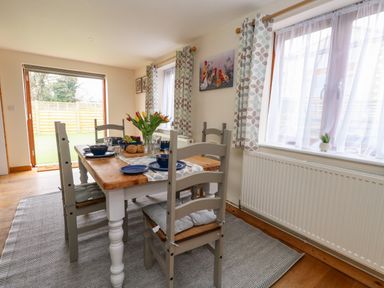 A dining room with a wooden table set with blue dishes and grey chairs near windows with patterned curtains at Little Dairy House near Swanage