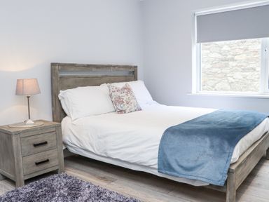 A bedroom with a wooden bed frame white bedding a blue blanket a floral pillow a wooden nightstand with a lamp and a window with a roller blind at Daly's Place in Ballyporeen County Tipperary