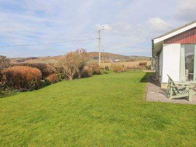A garden with grass and bushes next to a white house with chairs on the patio at Seafield in Rhydwyn near Cemaes Bay