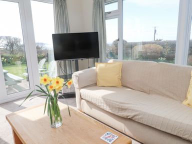 A living room with a beige sofa with yellow cushions a wooden coffee table with flowers and a TV near large windows at Seafield in Rhydwyn near Cemaes Bay
