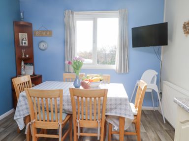 A dining room with a table and six wooden chairs a window with curtains a wall clock a wall mounted TV and a high chair at Seafield in Rhydwyn near Cemaes Bay