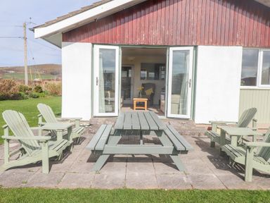 An outdoor patio with wooden chairs and a picnic table in front of a house with open glass doors at Seafield in Rhydwyn near Cemaes Bay