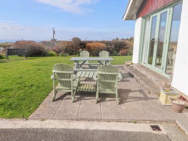 A patio with green wooden chairs and tables next to a house with glass doors and a garden with a bench at Seafield in Rhydwyn near Cemaes Bay