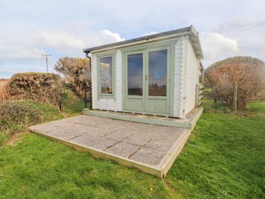 A small wooden shed with green windows and doors on a stone patio surrounded by grass at Seafield in Rhydwyn near Cemaes Bay