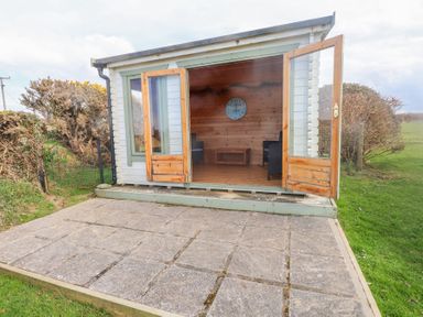 An outdoor wooden cabin with open double doors overlooking a paved patio and grassy area at Seafield in Rhydwyn near Cemaes Bay