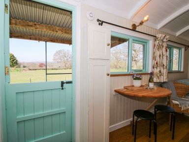A room with a turquoise half door opening to a field an indoor wooden table with two stools a wicker chair and windows with floral curtains at Gilberry Shepherds Hut Wall under Heywood near Church Stretton