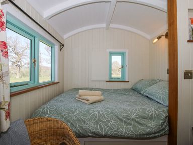 A small bedroom with a bed covered in green floral bedding folded towels on the bed a wicker chair and blue framed windows at Gilberry Shepherds Hut Wall under Heywood near Church Stretton