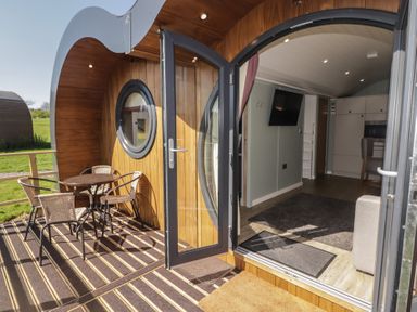 An outdoor wooden deck with a round table and four chairs leading to an open door showing a living area with a wall-mounted TV and kitchen at The Hazel Pod in Cowbridge