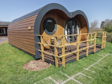 A wooden pod with circular and arched doors and a small deck with chairs and table on grass at The Hazel Pod in Cowbridge