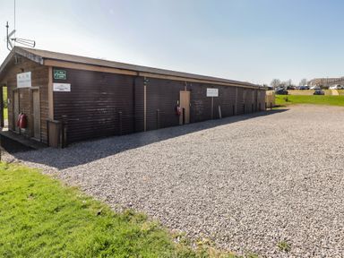 A long wooden cabin building next to a gravel area with parked cars in the background at The Hazel Pod in Cowbridge