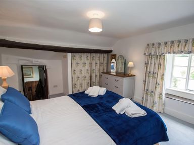 A bedroom with a bed covered by blue pillows and blanket a dresser with two trio mirrors window with floral curtains and a lamp at The Cragg in Hawkshead