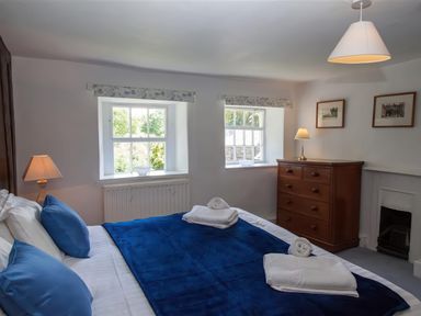 A bedroom with a double bed with blue pillows and blanket two windows wooden chest of drawers and lamps at The Cragg in Hawkshead