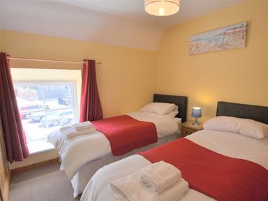 A bedroom with two single beds red bed runners white bedding a window with red curtains and a small bedside table with a lamp at Sunset Lodge in Fortuneswell