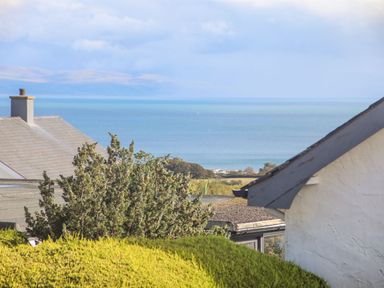 A view of a sea with rooftops, trees, and shrubs in the foreground at Trem Ynys 3 bed in Abersoch