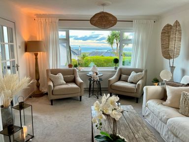A living room with two beige armchairs white sofa wooden coffee table and large window with sea view at Trem Ynys 3 bed in Abersoch