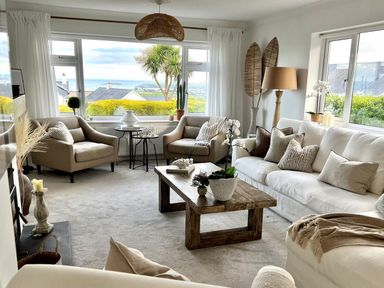 A living room with armchairs sofa coffee table and windows showing a garden and sea view at Trem Ynys 3 bed in Abersoch