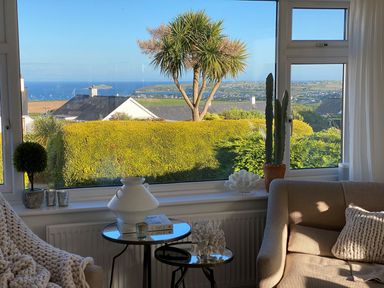 A living room with a beige sofa a knitted throw a window view of bushes trees and the sea at Trem Ynys (3 bed) in Abersoch