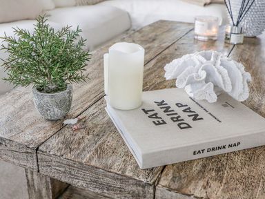 A wooden coffee table with a book titled Eat Drink Nap a white candle a small potted plant and a white decorative coral at Trem Ynys (3 bed) in Abersoch