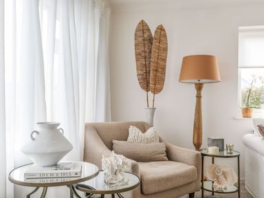 A beige armchair with cushions next to two glass and metal side tables with decorative items and a wooden floor lamp in a living room at Trem Ynys (3 bed) in Abersoch