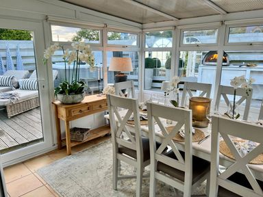 A dining area with a wooden table and white chairs next to a side table with a lamp and flowers at Trem Ynys (3 bed) in Abersoch