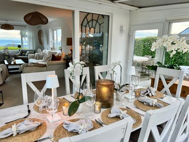 A dining table set with placemats, napkins, wine glasses, candles, and white orchids next to a living room with sofas at Trem Ynys 3 bed in Abersoch