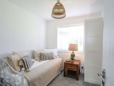 A small bedroom with a white metal daybed with pillows and a wooden bedside table with a lamp and decor at Trem Ynys (3 bed) in Abersoch