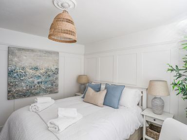 A bedroom with a bed with white bedding and folded towels on it with a large painting on the wall and bedside tables with lamps at Trem Ynys 3 bed in Abersoch