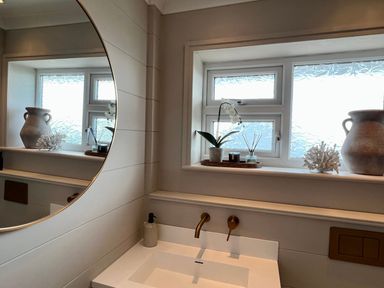 A bathroom sink with wall-mounted faucet and round mirror next to a window with decorative items on the sill at Trem Ynys 3 bed in Abersoch