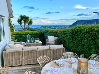 An outdoor patio with wicker furniture and a dining table set with glasses and napkins overlooking a garden and distant hills at Trem Ynys 3 bed in Abersoch