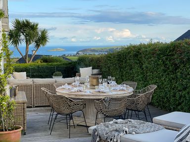 An outdoor patio with a round dining table set with glassware and napkins surrounded by wicker chairs and a wicker sofa with cushions overlooking a coastal view at Trem Ynys 3 bed in Abersoch