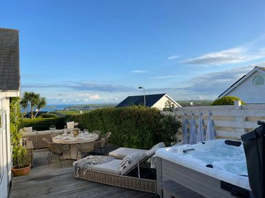 An outdoor patio with wicker dining table and chairs lounge chairs and a hot tub with a view of houses and the sea at Trem Ynys 3 bed in Abersoch