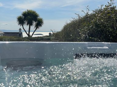 A bubbling hot tub outside with a view of palm trees bushes rooftops and water beyond at Trem Ynys 3 bed in Abersoch