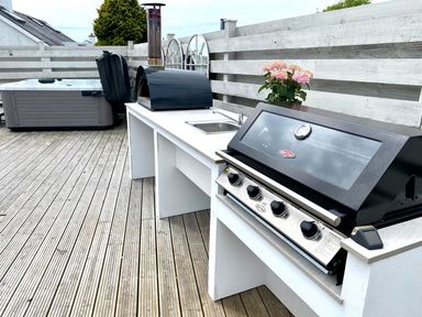 An outdoor deck with a hot tub a pizza oven a sink and a barbecue grill with a wooden fence behind at Trem Ynys in Abersoch