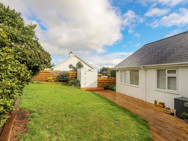 A backyard with green grass a white shed wooden fence and a house with a wooden deck at Trem Ynys in Abersoch