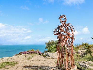 A metal sculpture of two people embracing on a rocky area with ocean and blue sky in the background at Trem Ynys (3 bed) in Abersoch