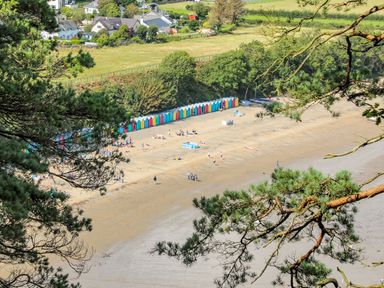A beach with colorful beach huts and people on the sand near houses and green fields at Trem Ynys (3 bed) in Abersoch