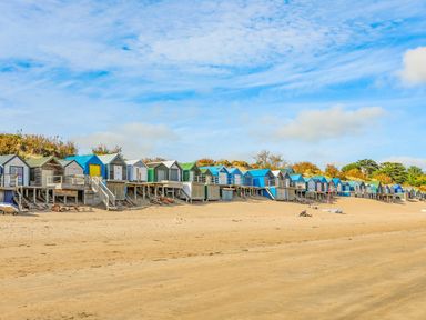 A row of colorful beach huts elevated on wooden stilts along a sandy beach under a blue sky at Trem Ynys 3 bed in Abersoch