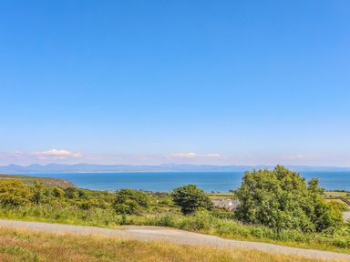 A coastal landscape with grassy fields trees and a road with the sea and distant mountains in the background at Trem Ynys 3 bed in Abersoch
