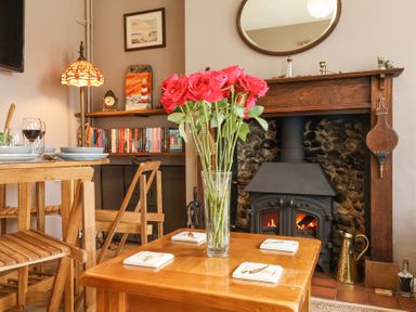 A room with a wooden table holding a vase of pink roses near a fireplace with a wood stove at 1 Rumsby Cottages in Gisleham near Kessingland