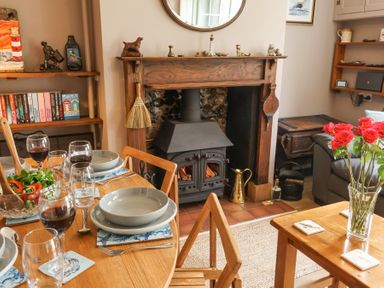 A dining area with a wooden table set for four with bowls glasses and wine near a wood burning stove and flowers in a vase at 1 Rumsby Cottages Gisleham near Kessingland
