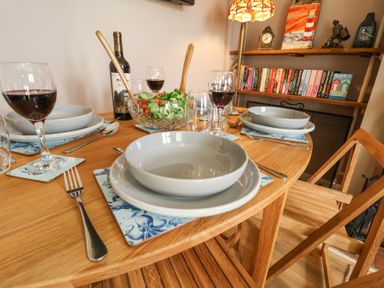 A dining table set with bowls plates wine glasses a salad bowl and a bottle of wine with a bookshelf in the background at 1 Rumsby Cottages Gisleham near Kessingland