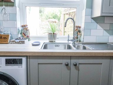 A kitchen sink area with a dual-basin stainless steel sink a chrome faucet a potted plant on the windowsill two decorative jars white tiled backsplash and light grey cabinets at 1 Rumsby Cottages Gisleham near Kessingland