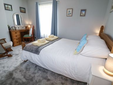 A bedroom with a bed with white bedding towels on a grey throw a wooden dressing table with a mirror a chair and framed pictures on the walls at 1 Rumsby Cottages Gisleham near Kessingland