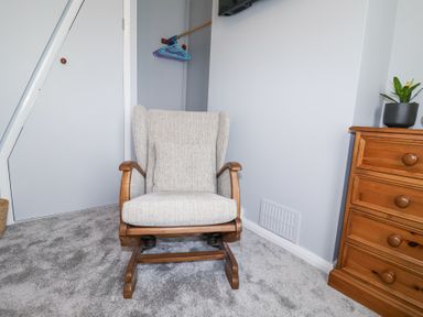 A room with a beige rocking chair a wooden dresser with a plant a door and gray carpet at 1 Rumsby Cottages Gisleham near Kessingland