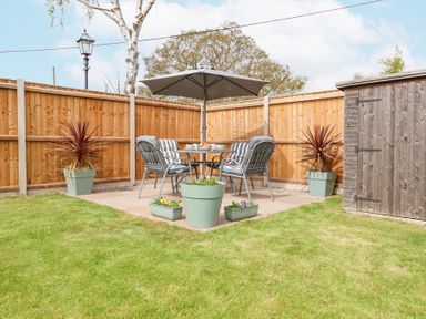 A backyard patio with four chairs around a table under an umbrella next to a wooden shed and fenced yard at 1 Rumsby Cottages Gisleham near Kessingland