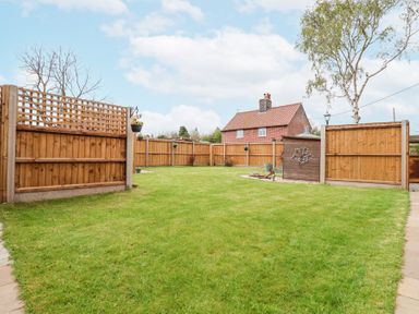 A fenced backyard with grass a wooden shed and a brick house in the background at 1 Rumsby Cottages Gisleham near Kessingland
