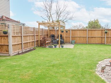 A backyard with a wooden pergola, two chairs, flower pots, and a grass lawn at 1 Rumsby Cottages Gisleham near Kessingland