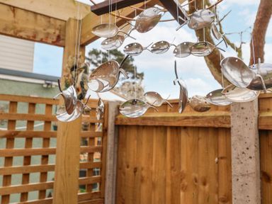 An outdoor wooden pergola with a metallic fish wind chime hanging in front of a wooden fence and trees at 1 Rumsby Cottages in Gisleham near Kessingland