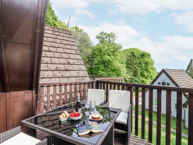 A balcony with a table set with food drinks and books overlooking trees and neighboring houses at 23 Valley Lodge in Gunnislake