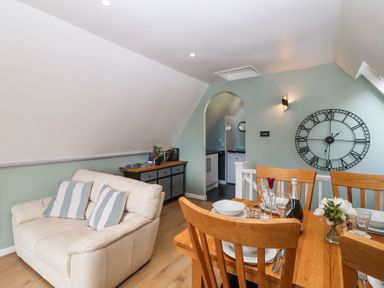 A dining table with wooden chairs set with plates and glasses in a room with a cream sofa and a large wall clock at 23 Valley Lodge in Gunnislake